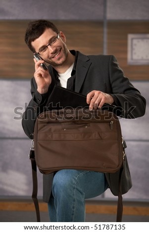 Happy office worker talking on mobilephone, taking computer out of laptop bag, smiling.