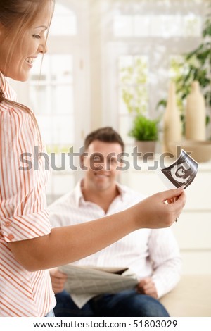 Smiling woman looking at baby\'s ultrasound picture held in hand, man smiling in background at home.