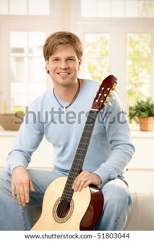 Portrait of young man sitting in living room with guitar, smiling at camera.