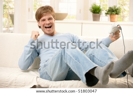 Happy guy playing computer game, holding controller, laughing on living room sofa.