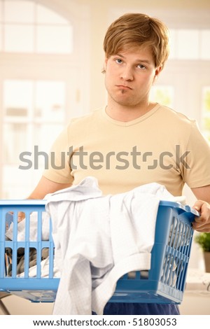 Young troubled guy standing holding washing basket, bored of housework, looking at camera.