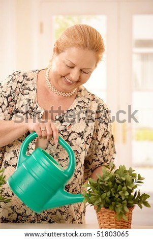 Smiling senior blond woman watering green potted plant from sprinkling can.