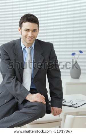 Portrait of businessman wearing grey suit and blue shirt, sitting on office desk, smiling.