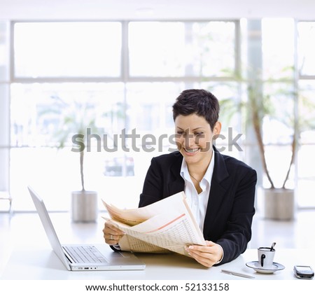 Happy businesswoman sitting at table in office lobby, reading newspaper, using laptop computer.
