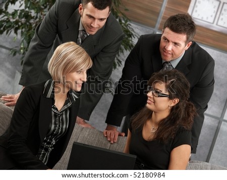 Young businesswomen sitting at couch in office talking with businessmen standing behind, smiling.