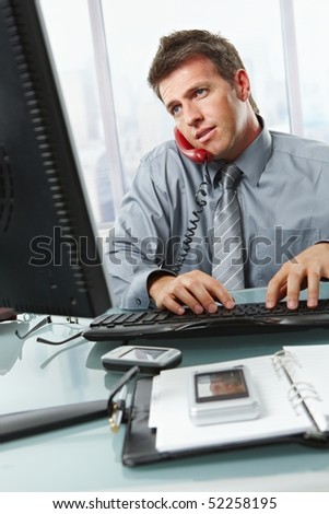 Businessman at office desk busy with typing on keyboard, talking on landline phone and looking at screen.