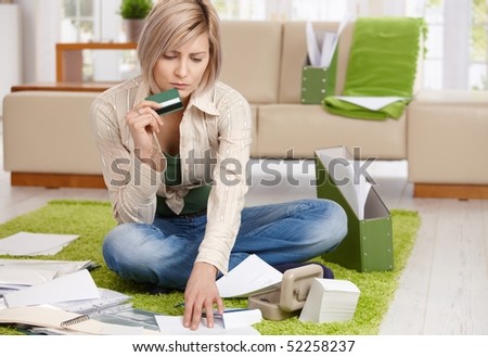 Worried woman checking documents, holding credit card, sitting on floor with crossed legs at home.