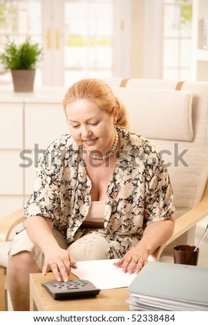 Blonde senior woman sitting in armchair in living room, doing financial calculation on paper, using calculator, smiling.