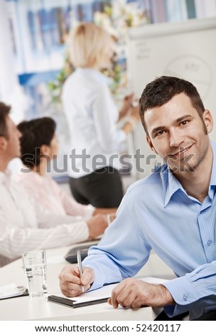 Happy businessman sitting at table in office writing notes on business meeting, looking at camera, smiling.