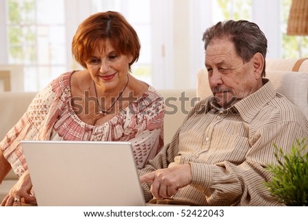 Elderly couple using laptop computer at home, looking at screen, smiling.