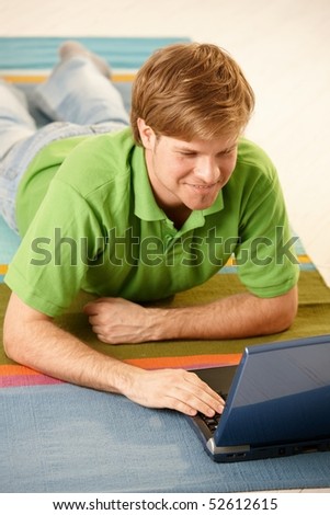 Man lying on colorful carpet, using laptop computer browsing Internet, looking at screen, smiling.