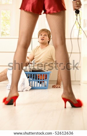 Woman wearing netted tights, sexy red baby-doll and high heels slippers holding lash forcing young man with laundry basket to do housework.