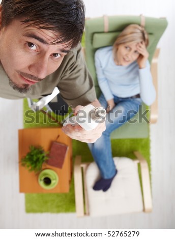 In overhead view man in focus changing light bulb standing on ladder, woman in background sitting in armchair, holding head, smiling.