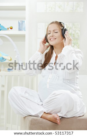 Pregnant woman sitting besides new crib in children's room, listening music.