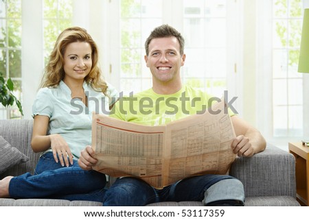 Happy couple reading newspaper together on couch at home, looking at camera, smiling.