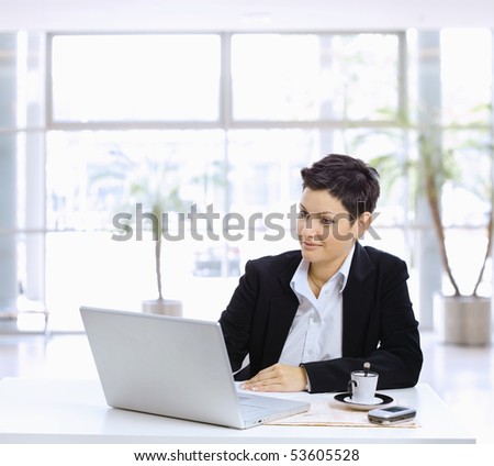 Businesswoman sitting at table in office lobby, using laptop computer, looking at screen.