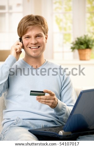 Smiling man talking on mobile phone, holding creditcard, sitting on couch with laptop computer, looking at camera