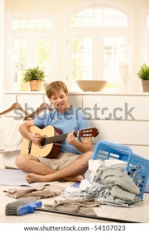 Lazy young guy playing guitar sitting on floor instead of washing laundry or sweeping.