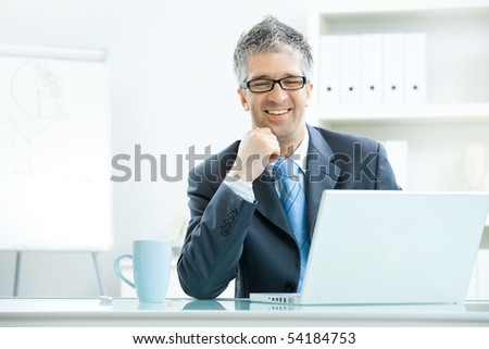 Businessman with grey hair, wearing grey suit and glasses thinking over laptop computer, sitting at desk in bright, modern office, leaning on hand, smiling.
