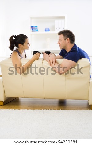 Young couple drinking coffee at home sitting on couch. Smiling and looking at each other.