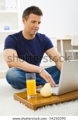 Casual young man using laptop computer at home, sitting at floor, looking at screen.