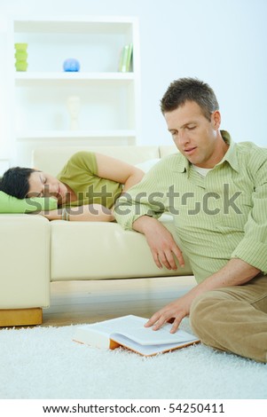 Young couple resting at home. Man reading book on floor, woman sleeping on couch.