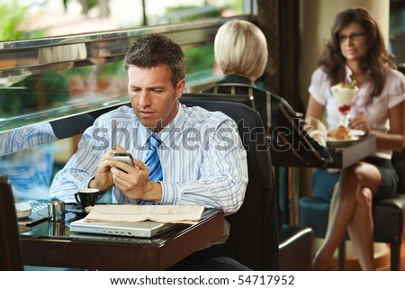 Businessman sitting at table in cafe using mobile phone. Young women having sweets in the background.