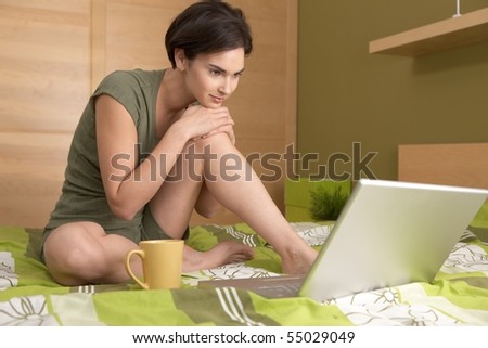 Smiling woman looking at laptop sitting on bed with coffee mug.