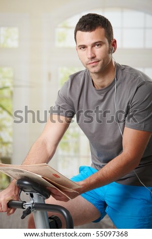 Man sitting on stationary bike at home, reading newspaper and listening music.