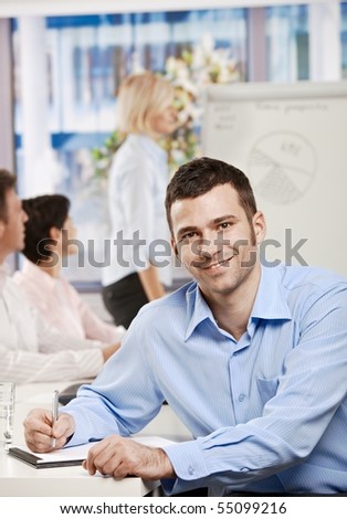 Happy businessman sitting at table in office writing notes on business meeting, looking at camera, smiling.?