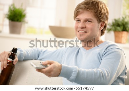 Smiling guy changing channels with remote control, sitting at home with beer in hand.?