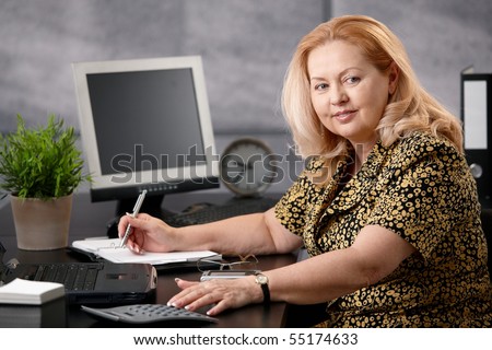 Senior woman sitting at office desk working, writing notes to personal organizer, smiling.?