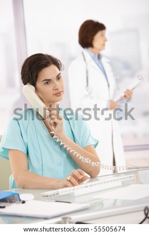 Assistant sitting at desk taking phone call using computer, medical doctor standing in background.