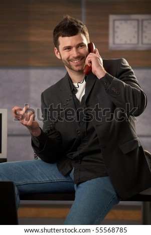 Happy businessman laughing making landline phone call, gesturing with hand, sitting on office desk.