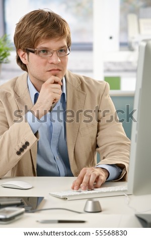 Young businessman sitting at office desk, looking at screen thinking.