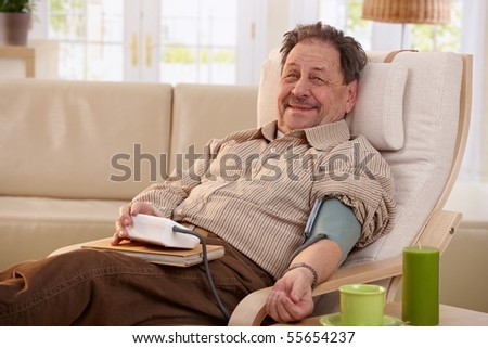 Elderly man sitting in armchair at home, measuring his blood pressure, smiling.