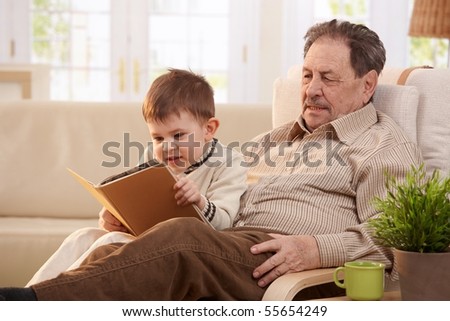 Grandfather sitting in armchair at home, holding his grandson who looking pictures in book.
