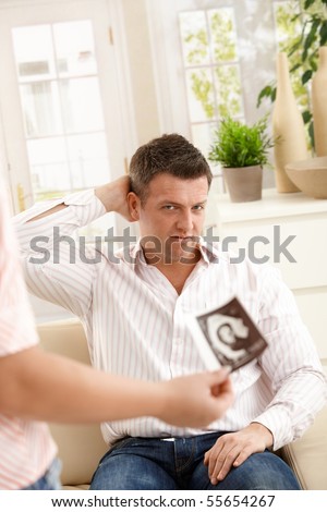 Man looking unbelieving at ultrasound picture of baby, holding his head sitting on couch.