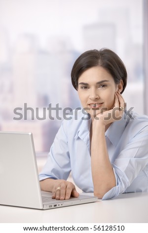 Portrait of mid adult woman sitting at office table with laptop computer, smiling at camera.