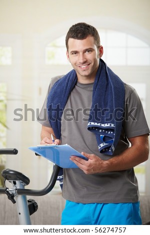 Man standing beside stationary bike after finishing exercise, making notes in training plan.