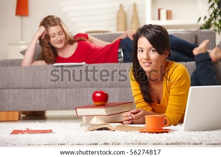 Teenage girls learning at home in living room with books and laptop, girl in front smiling at camera.
