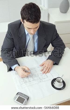 Architect wearing grey suit sitting at office desk, writing notes on floor plan. Overhead shot.