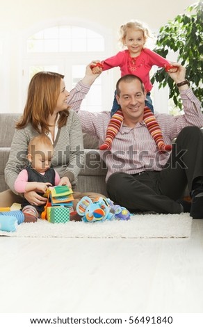 Happy family of four sitting at home on floor of living room, smiling girl sitting in daddy\'s neck.