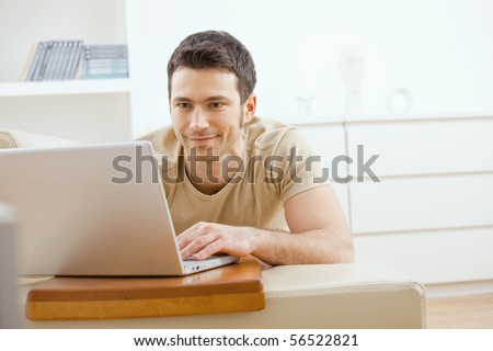 Happy young man laying on sofa at home using laptop computer, smiling.