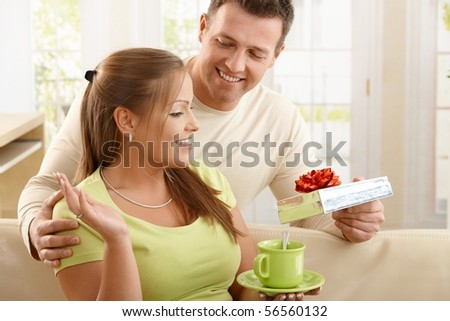 Woman sitting in living room drinking tea, smiling man surprising happy woman with present.