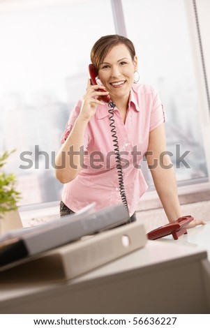 Happy office worker talking on phone, leaning on office desk, smiling.