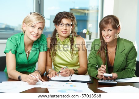 Young  businesswomen sitting at table in meeting room, discussing charts on table, looking at camera, smiling.