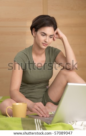 Woman using laptop computer in bed, sitting with coffee mug, smiling.?