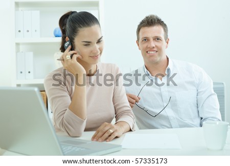 Couple working on laptop computer at home office, happy, smiling. Woman calling on mobile phone.