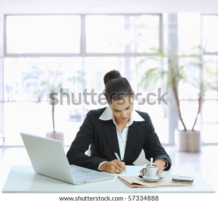 Businesswoman sitting at table in office lobby, writing note on paper.